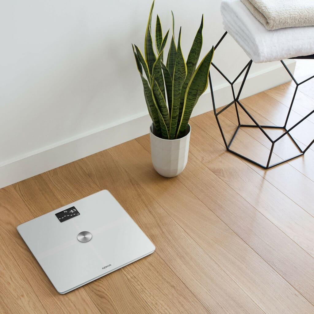 a white withings smart scale is on a floor next to a snake plant