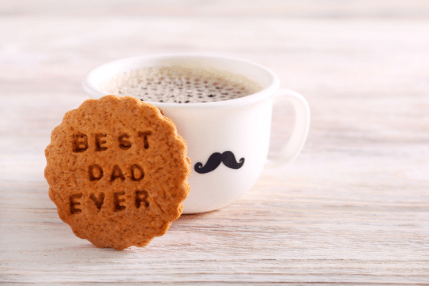 A close-up photo of a mug filled with steaming coffee and a delicious cookie with the words "Best Dad Ever" written on it, a perfect treat to celebrate Father's Day with PostFromUS.