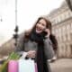 A cheerful woman answering a mobile phone call with her left hand and carrying paper shopping bags on her right arm.