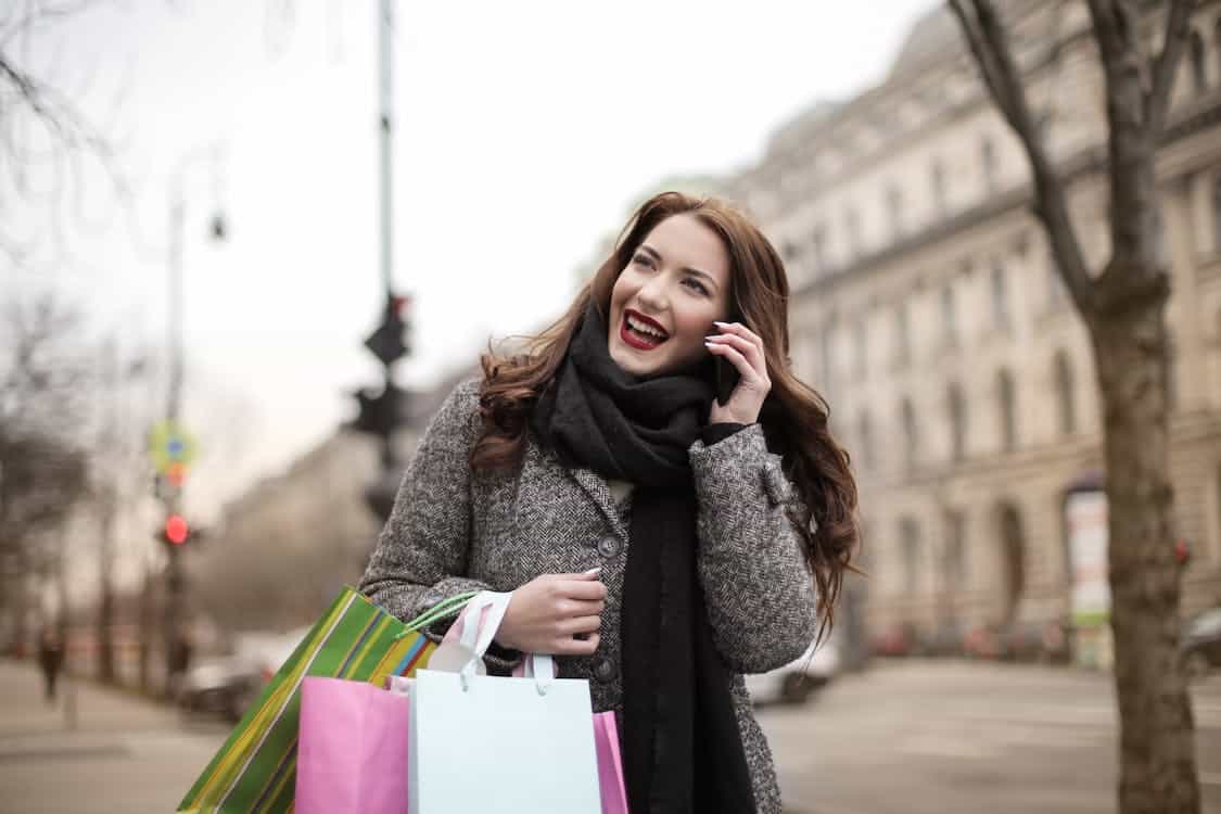 A cheerful woman answering a mobile phone call with her left hand and carrying paper shopping bags on her right arm.