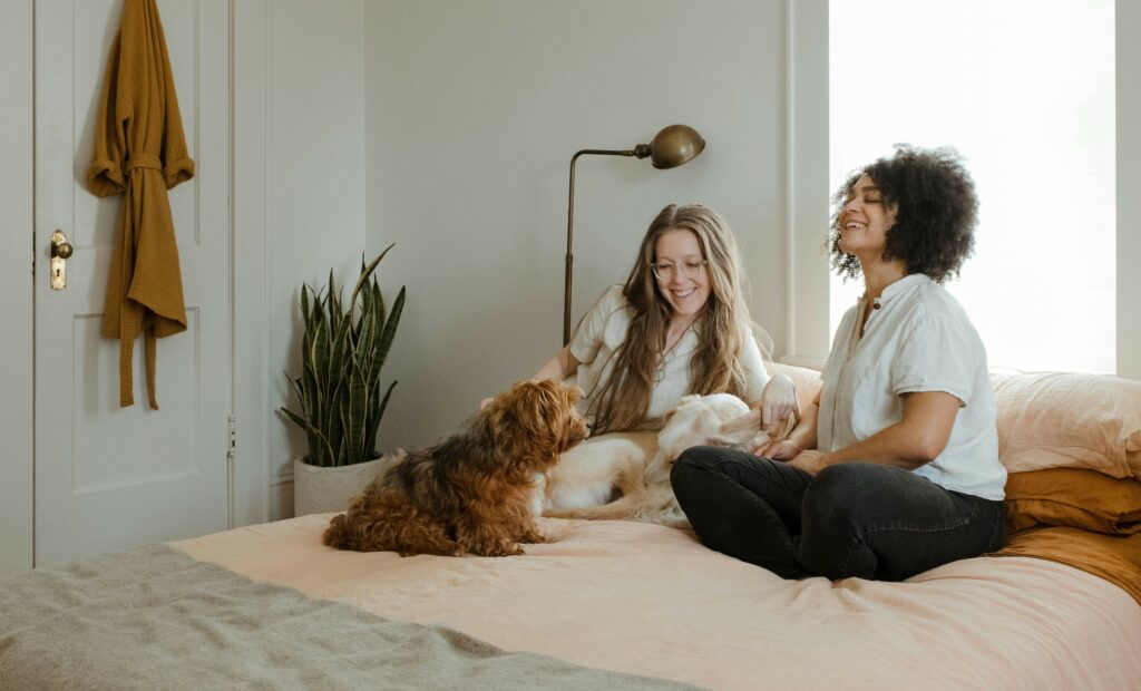 Two girls sit on a bed with a dog in front of them.