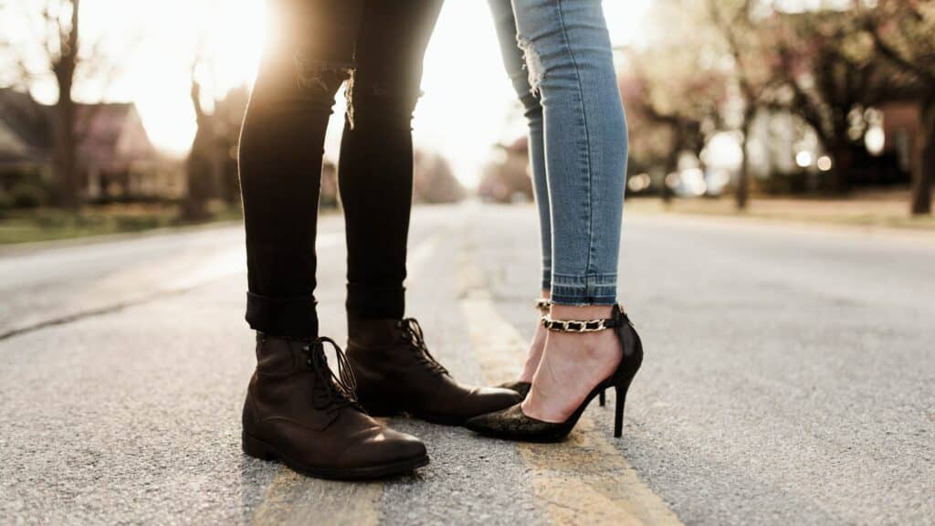 A man and a woman meet in the middle of a road, the camera is focused on their feet. He is wearing black shoes, she is wearing black heels. 