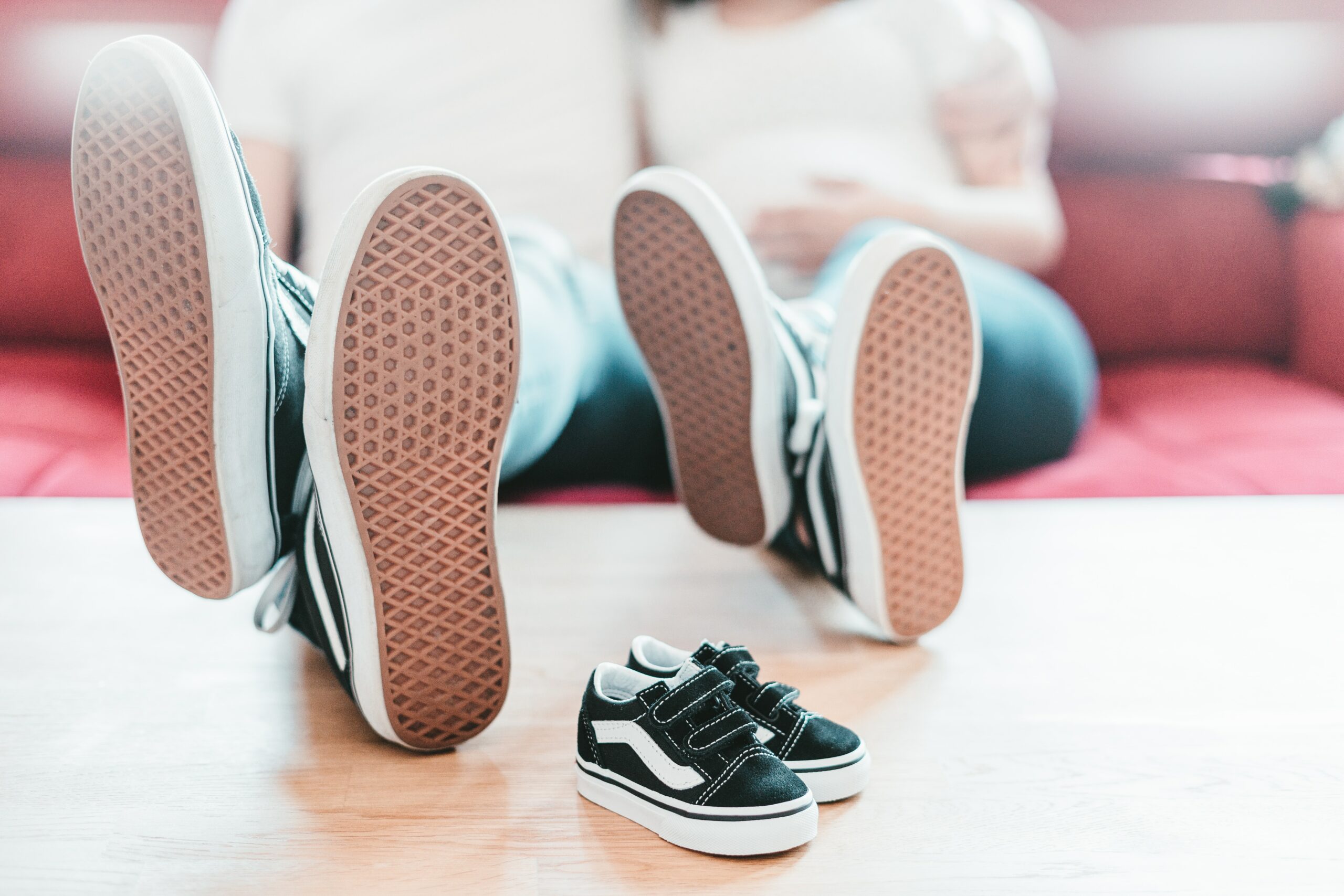 Two people have their feet up on a coffee table with their shoes on. An empty pair of baby shoes is also on the table.