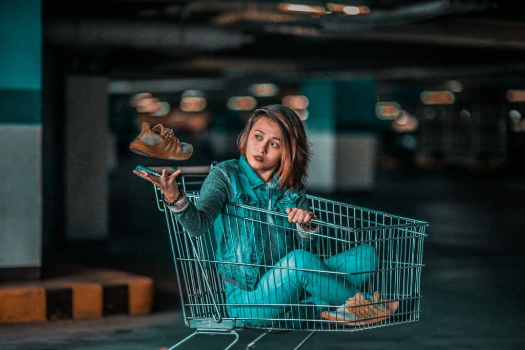 A woman is sitting in a shopping cart in a parking garage, holding a shoe in her hand. She has a look of contemplation.