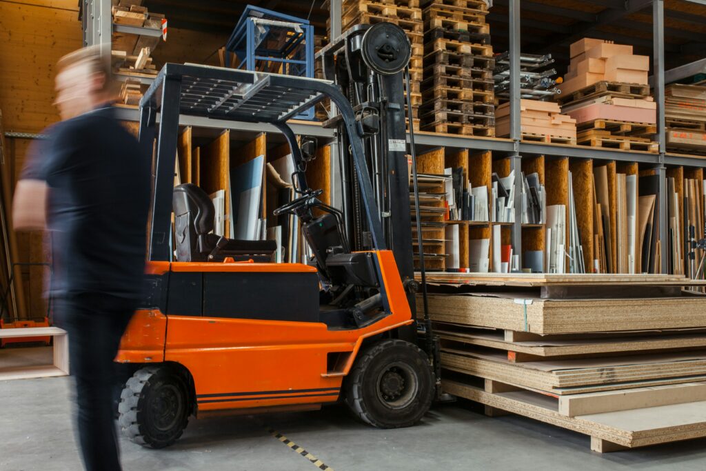 A warehouse worker walks past a forklift, which is parked in front of fully stocked shelves.