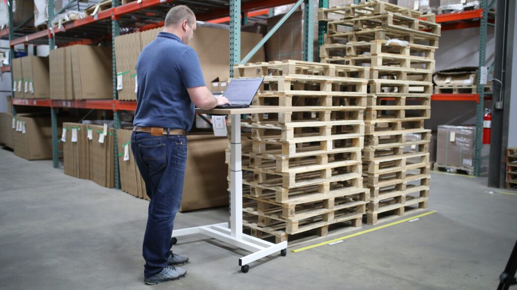 A man in a warehouse is working on a laptop near some wood palettes. 