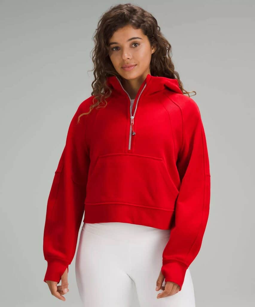 a woman is standing in front of a grey background wearing white leggings and a red half zip thumb hole hoodie. her hair is brown and curly and she's smiling. 