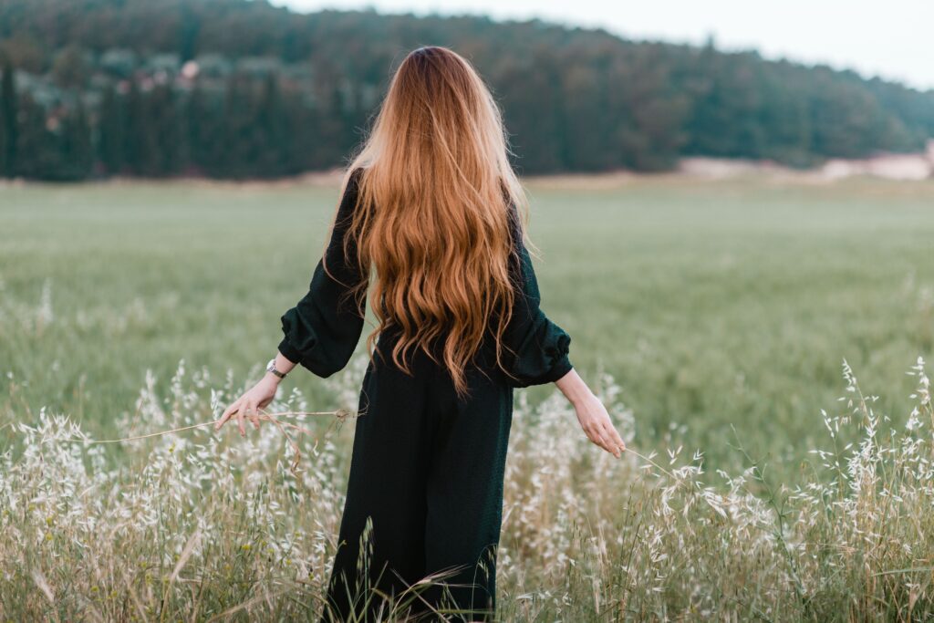 A woman with long red hair is wearing a black dress with pockets in a field with her arms out beside her.