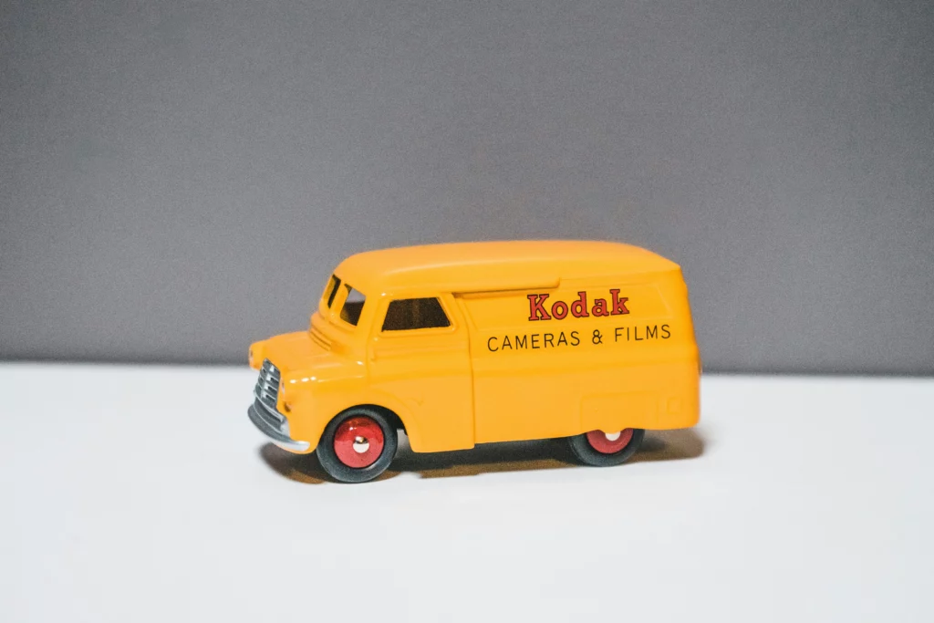 a yellow kodak van from a model car collection on a white table with a grey background.