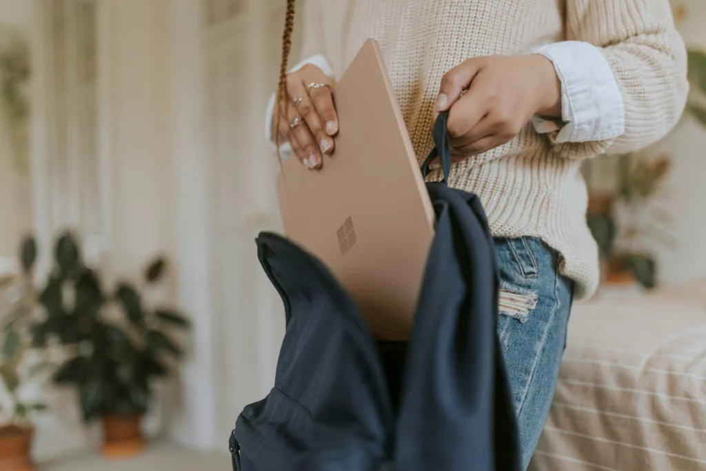 Someone is wearing a white sweater, putting a rose gold microsoft surface pro laptop from america in their blue backpack