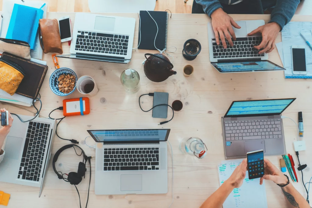 An overhead view of many gadgets on a desk, where people are working out of frame. There are Macbooks, headphones, and several American gift ideas there.