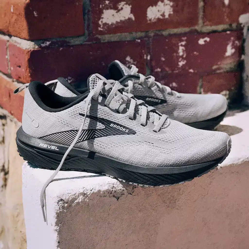 Brooks Running, que tiene diferentes tallas de calzado en Estados Unidos que en la UE o China. Son de color blanco o gris y están colocados en un escalón de hormigón frente a una pared de ladrillos.