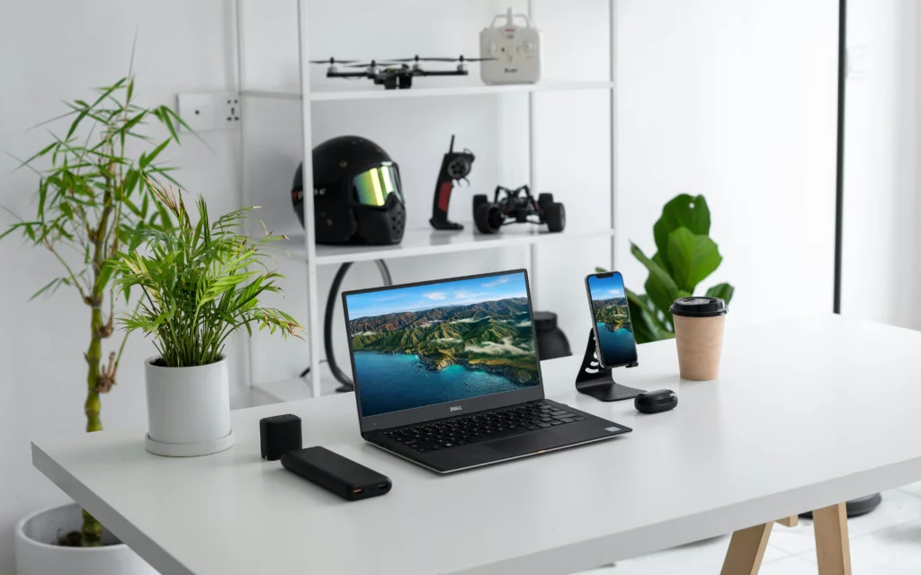 Un escritorio con un ordenador portátil y varios aparatos electrónicos. Las paredes son blancas y hay una planta junto al escritorio y encima de él.