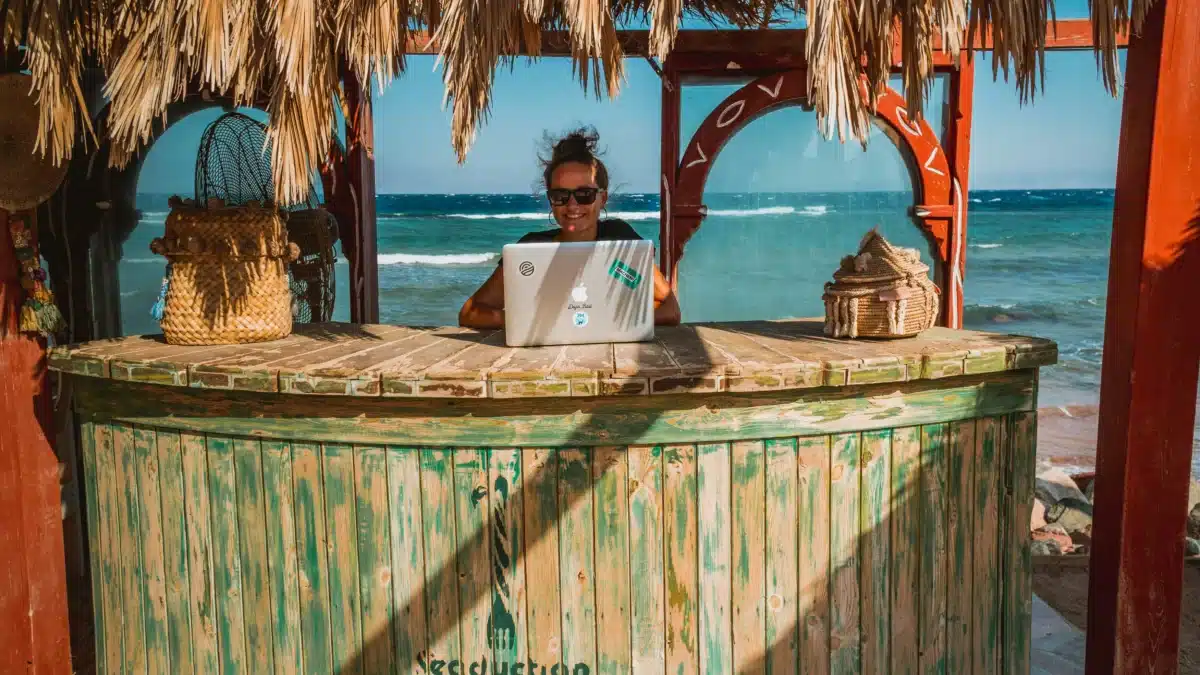 A woman is in a straw hut on a sunny beach, smiling as she uses her laptop to get a free mailing address to deliver all her favorite American goods to.