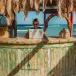 A woman is in a straw hut on a sunny beach, smiling as she uses her laptop to get a free mailing address to deliver all her favorite American goods to.