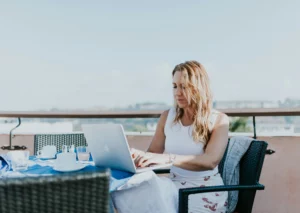 A blonde woman is sitting in a white outfit on a balcony. There is an outdoor dining set. She has a laptop on the table. She is learning how to leverage a free mailing address.