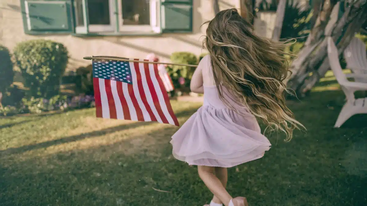 A little girl in a dress is running outside of a house on grass with an American flag blowing in the wind from her arms. American gift ideas are loved by most children.
