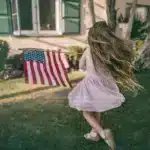 A little girl in a dress is running outside of a house on grass with an American flag blowing in the wind from her arms. American gift ideas are loved by most children.