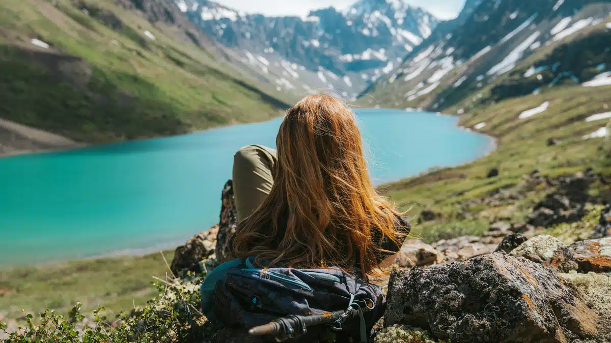 A girl is sitting against a rock facing a mountain and a river after receiving her camping gear. Shipping in Alaska is easy!