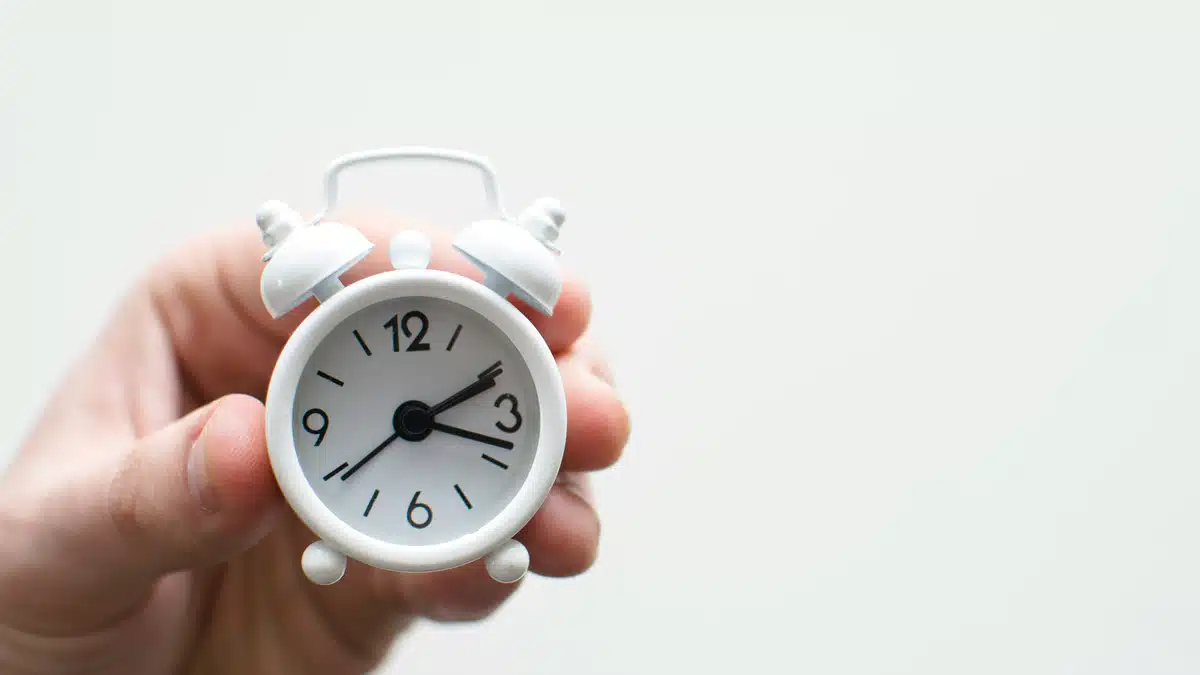 Someone is holding a little white alarm clock. You can only see their hand.